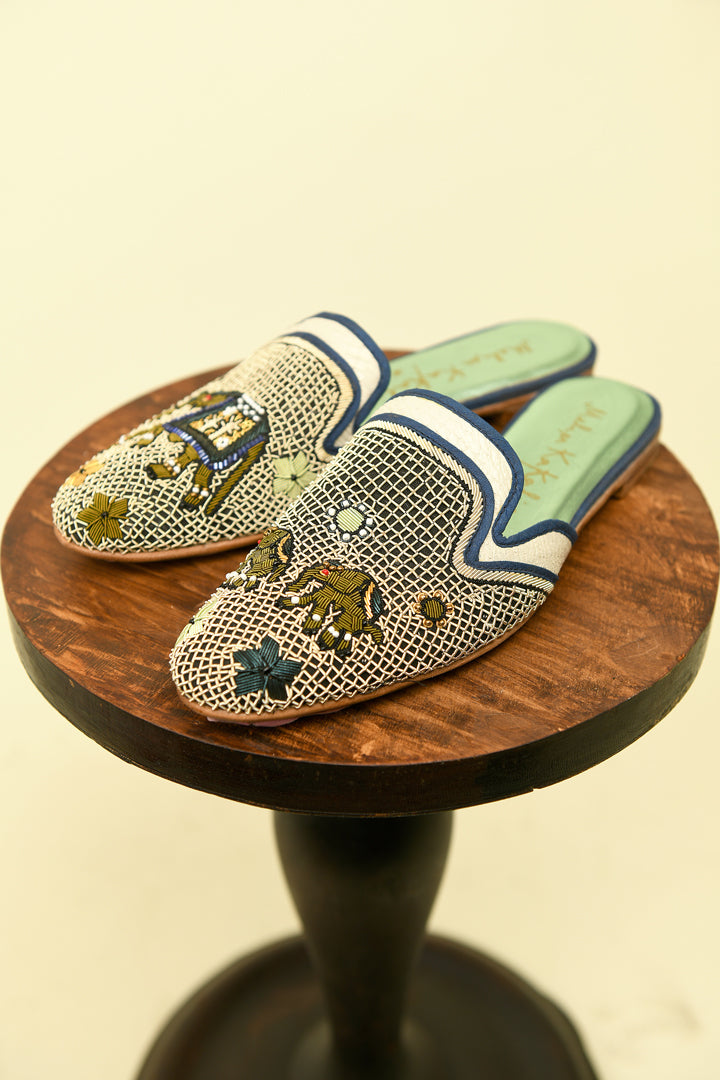 Embroidered slip on shoes with a mother elephant on the left shoe and two baby elephants on the right shoe.  Close up of shoes from a side angle on a wooden pedestal  on a cream background.